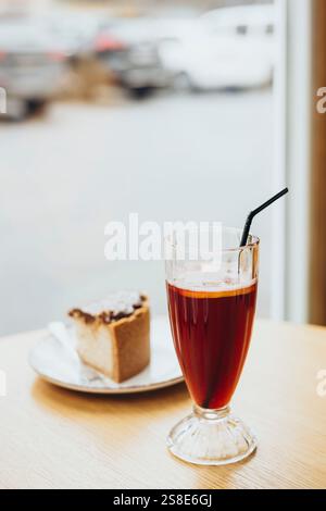 Un verre de thé glacé avec une paille et une tranche de gâteau sur une table en bois près d'une fenêtre, offrant une ambiance détendue dans un cadre de café confortable Banque D'Images
