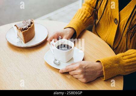 Une personne dans un pull jaune s'assoit à une table en bois, savourant une tasse de café à côté d'une tranche de gâteau la scène évoque chaleur et détente Banque D'Images
