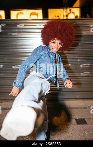 Femme joyeuse descendante africaine aux cheveux afro rouges distinctifs, portant des lunettes et une tenue en denim, danse de manière ludique dans un environnement urbain nocturne Banque D'Images