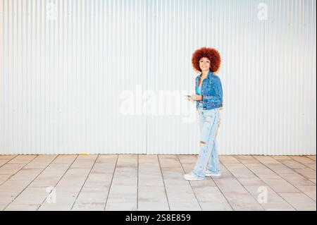 Femme de descendance africaine avec des cheveux afro vibrants, portant une veste en denim et un Jean déchiré, se tient souriant sur un fond rayé, tenant un chic Banque D'Images