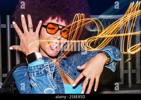 La femme descendante africaine aux cheveux afro met en valeur son côté ludique, claquant et tirant sa langue avec des lunettes de soleil teintées d'orange la nuit. Lumière Banque D'Images