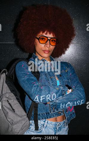 Femme d'ascendance africaine avec une coiffure afro volumineuse portant des lunettes de soleil orange et une veste en denim, posant en toute confiance sur un fond sombre avec Banque D'Images