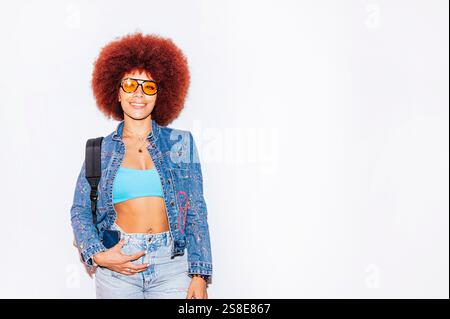 Femme de descendance africaine avec une chevelure afro vibrante, souriante tout en portant des lunettes de soleil orange et une veste en denim, debout contre un backgrou blanc Uni Banque D'Images