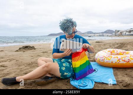 Un homme transgenre aux cheveux bleus assis sur une plage, mettant une tablette dans un sac de fierté arc-en-ciel. Il est habillé d'une chemise bleue et d'un short fleuri, symb Banque D'Images