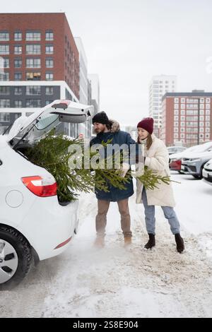 Un couple charge joyeusement un sapin de Noël dans un coffre de voiture dans un cadre urbain enneigé, se préparant pour la saison des fêtes, ils sont regroupés dans l'hiver chaud cl Banque D'Images