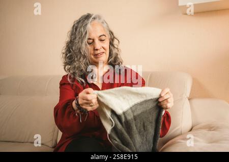 Une femme en pull rouge, aux cheveux gris, est assise sur un canapé beige tricotant une pièce grise et blanche. Elle est souriante et semble détendue dans sa vie confortable Banque D'Images