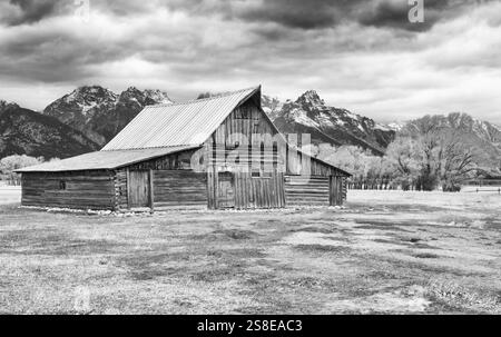 Une image en noir et blanc capturant une ancienne grange en bois avec la majestueuse gamme Grand Teton en arrière-plan sous un ciel nuageux. Mormon Row dans le Wyoming, Banque D'Images
