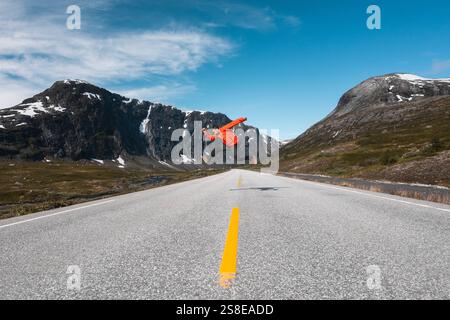 Un voyageur dans une veste rouge saute en plein air sur une route de montagne ouverte en Norvège encadrée par des sommets enneigés spectaculaires et un ciel bleu vibrant, cette scène RA Banque D'Images