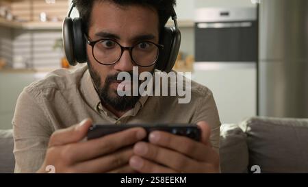 Choqué étonné homme arabe indien regarder la vidéo streaming sport en ligne match addict des médias sociaux homme concentré dans les écouteurs regardant le téléphone mobile Banque D'Images