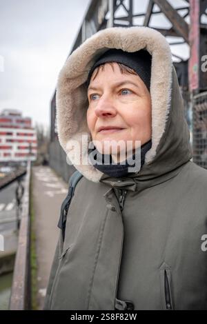 Une femme blanche mature portant une veste à capuche kaki se tient sur un pont et regarde l'horizon en hiver Banque D'Images