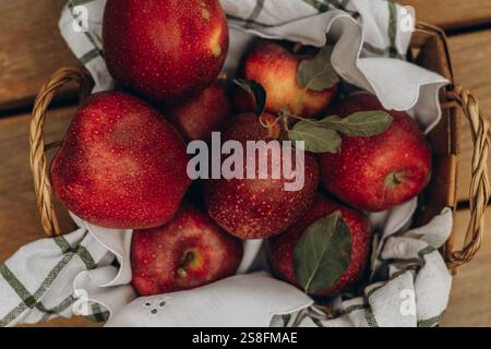 Vue de dessus du panier rustique rempli de pommes juteuses et rouges, accentuées par des feuilles vertes et un tissu rayé, présentant une scène fraîche et organique. Banque D'Images