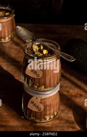 Une mousse d'avocat au chocolat végétalien magnifiquement stylisée garnie de pistaches, servie dans un bocal en verre avec une étiquette d'amour, positionnée sur une surface en bois foncé Banque D'Images