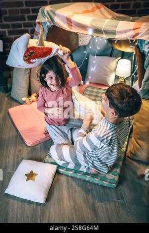 Joyeux garçon et fille avec pyjama riant et jouant dans la bataille de coussin à la maison devant la tente Banque D'Images