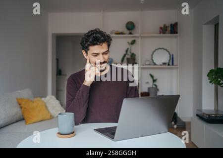 Jeune homme beau moyen-oriental regardant à l'aide d'un ordinateur portable dans sa maison. Banque D'Images