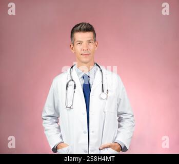 Beau jeune médecin en manteau blanc, stéthoscope autour du cou, cravate et chemise, mains dans les poches de manteau, regardant la caméra. Fond rose Banque D'Images