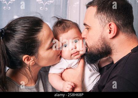 Un père aimant tient son fils bébé solidement dans ses bras tandis que la mère se tient proche, et les deux parents embrassent doucement les joues de leur enfant contre A. Banque D'Images