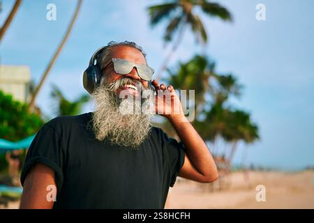 Homme senior avec la barbe grise dans des lunettes de soleil écoute de la musique sur la plage tropicale. Élégant, chic, il aime les vibrations estivales avec des écouteurs modernes. Ambiance détendue Banque D'Images