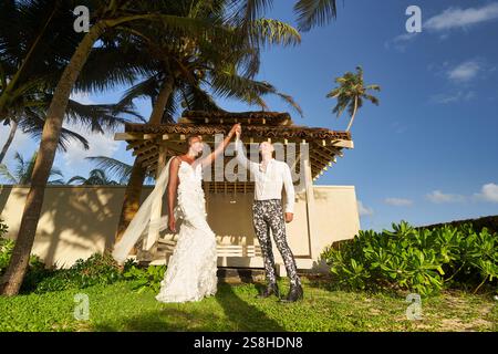 Mariage tropical avec mariée en robe blanche et marié LGBTQ en costume audacieux. Les couples fluides de genre ambigus dansent près de la villa. Journée ensoleillée, palmiers Banque D'Images