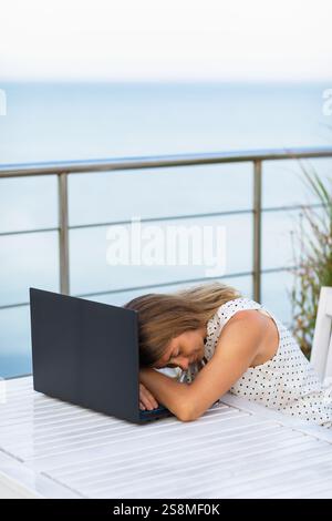 Femme penchée sur ordinateur portable se relaxant dans un café d'été avec vue sur la mer, concept sur le thème du repos du travail Banque D'Images