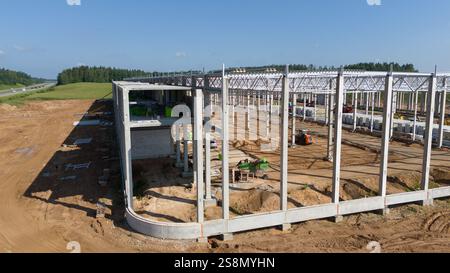 Vue aérienne d'un chantier de construction présentant une grande charpente de bâtiment. Le site est entouré de terres ouvertes et d'arbres, avec du matériel de construction vi Banque D'Images