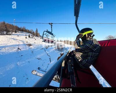 Un skieur s'assoit confortablement sur une remontée mécanique, profitant d'une vue panoramique sur les montagnes enneigées sous un ciel bleu clair. Une activité de plein air hivernale sereine dans un Banque D'Images