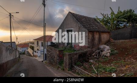 Une petite maison altérée sur le côté d'une route rurale en pente, avec des bananiers et des maisons modernes en arrière-plan. La douce lumière du soir améliore Banque D'Images