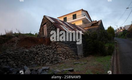 Une vieille maison en pierre avec un toit en métal rouillé contraste avec une maison moderne derrière elle. La structure rurale, construite à partir de pierres brutes, met en valeur le Banque D'Images