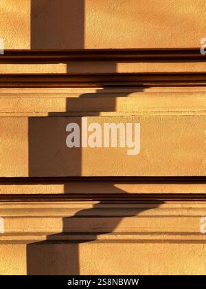 Une composition minimaliste mettant en valeur des ombres géométriques projetées sur un mur en stuc jaune texturé, illuminé par la lumière chaude du soleil. Banque D'Images