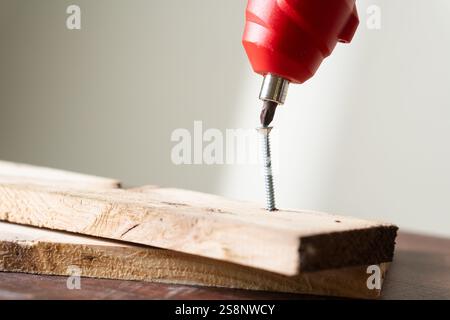 Gros plan d'un tournevis électrique rouge insérant une vis dans un morceau de bois. L'accent est mis sur le tournevis et la vis, avec des planches en bois dans le Banque D'Images