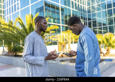 Deux hommes d'affaires sénégalais portant des vêtements traditionnels de dashiki utilisent des smartphones devant un immeuble de bureaux moderne, discutant du travail et de la technolo Banque D'Images