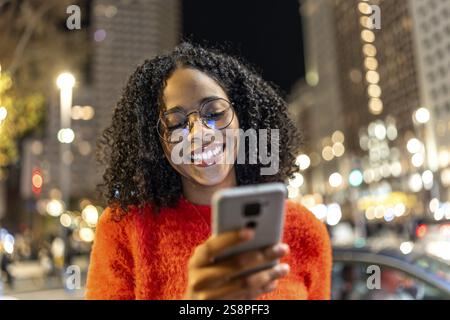 Femme souriante utilisant son smartphone tout en profitant de la vie nocturne urbaine animée, entourée par les lumières de la ville et une atmosphère animée Banque D'Images
