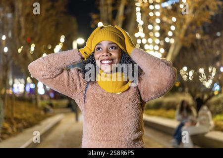 Jeune femme souriante portant des vêtements d'hiver ajustant son chapeau de laine jaune sous les lumières de noël dans un parc de la ville la nuit Banque D'Images