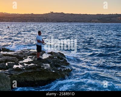 Jeune homme pêchant sur les rochers dans la mer au coucher du soleil Banque D'Images