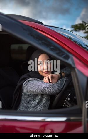 femme asiatique avec un hijab noir et un chemisier gris assis dans le siège du conducteur d'une voiture rouge, souriant légèrement tout en regardant soigneusement par la fenêtre Banque D'Images