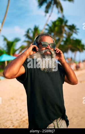 L'homme âgé avec la barbe grise aime la musique sur la plage ensoleillée. Lunettes de soleil et écouteurs haut de gamme. Ambiance relaxante dans un cadre tropical avec palmiers Banque D'Images