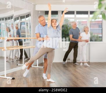 Femme âgée souriante portant des vêtements de sport dansant en couple et la formation ballet arabesque pose avec partenaire pendant le cours de groupe au salon de danse Banque D'Images