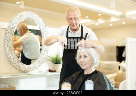 Le maître expérimenté de l'homme senior fait effectue la coupe de cheveux pour la femme mature avec des ciseaux Banque D'Images