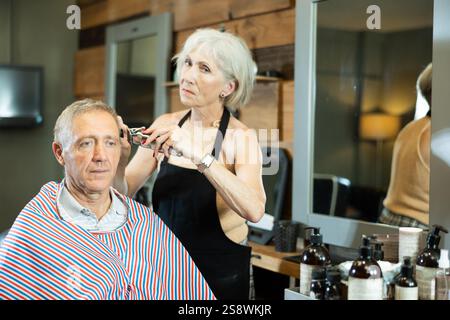 Le maître expérimenté de la femme mature fait effectue la coupe de cheveux pour l'homme âgé avec tondeuse Banque D'Images