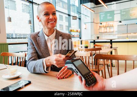 Joyeuse femme mature s'engageant dans Seamless faire un paiement avec carte de crédit au caissier dans Bright and Incoming Cafe. Interactions transactionnelles modernes et excellence du service à la clientèle. Photo de haute qualité Banque D'Images