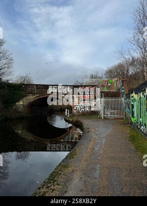 Un pont de pierre qui se reflète dans l'eau calme du canal Rochdale à Manchester, avec de l'art de rue dynamique et des graffitis le long du chemin de halage urbain. Banque D'Images