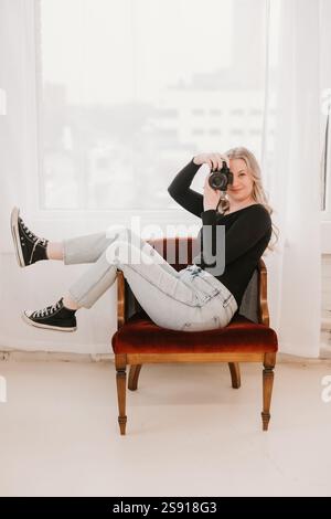 Woman playfully posing with a camera on a red chair by a window Banque D'Images