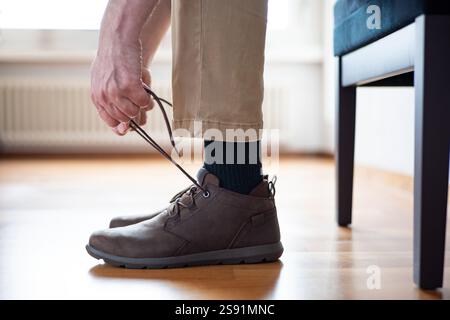Homme méconnaissable lacant ses chaussures semi-décontractées en cuir marron. Gros plan en angle bas, prise de vue intérieure, lumière naturelle. Banque D'Images