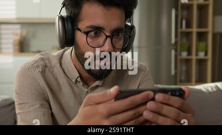 Choqué étonné homme arabe indien regarder la vidéo streaming sport en ligne match addict des médias sociaux homme concentré dans les écouteurs regardant le téléphone mobile Banque D'Images