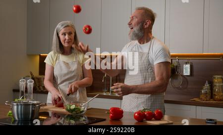 Caucasien famille heureuse couple d'âge moyen retraité dans la femme amoureuse cuisinant le petit déjeuner de nourriture dans la cuisine drôle vieil homme jonglant avec des tomates légumes frais Banque D'Images