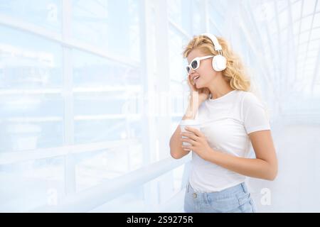 Belle jeune femme avec des lunettes de soleil et des écouteurs de musique, debout sur le pont avec une tasse de café à emporter et souriante Banque D'Images