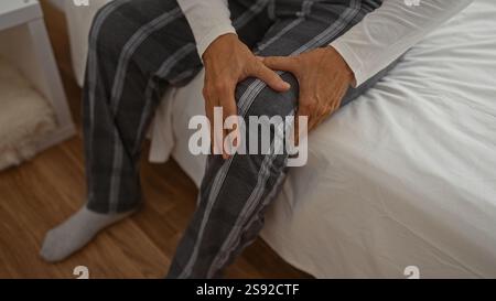 Homme tenant son genou avec les deux mains tout en étant assis sur un lit dans une chambre en pyjama Banque D'Images