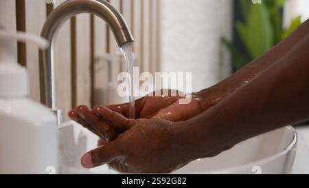 Gros plan homme afro-américain se lavant les mains sales avec de l'eau chaude savonneuse dans la salle de bain de l'hôtel en utilisant le désinfectant pour les mains hygiène lavage soin rincer les paumes dedans Banque D'Images
