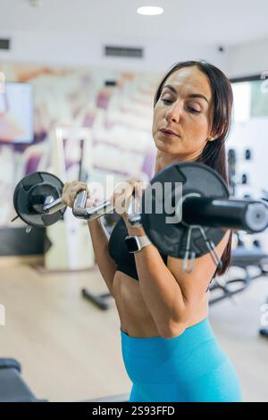 Une femme soulève des poids dans une salle de gym. Elle porte un haut noir et un pantalon bleu. La femme est concentrée sur son entraînement et elle est d'humeur déterminée Banque D'Images