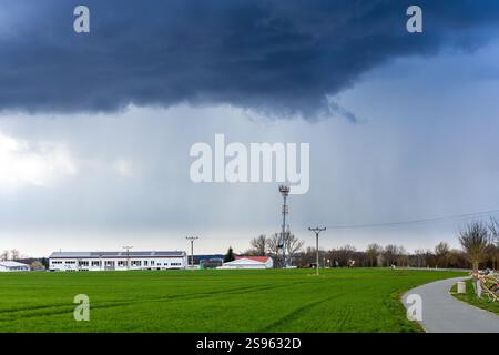 Nuages sombres sur un champ vert avec un chemin sinueux, des lignes électriques et une tour de communication près d'une zone rurale avec des bâtiments industriels blancs Banque D'Images
