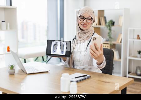 Médecin musulman féminin effectue un appel vidéo avec le patient, affichant une radiographie pulmonaire sur tablette. Stéthoscope professionnel d'âge moyen en cabinet médical. Banque D'Images